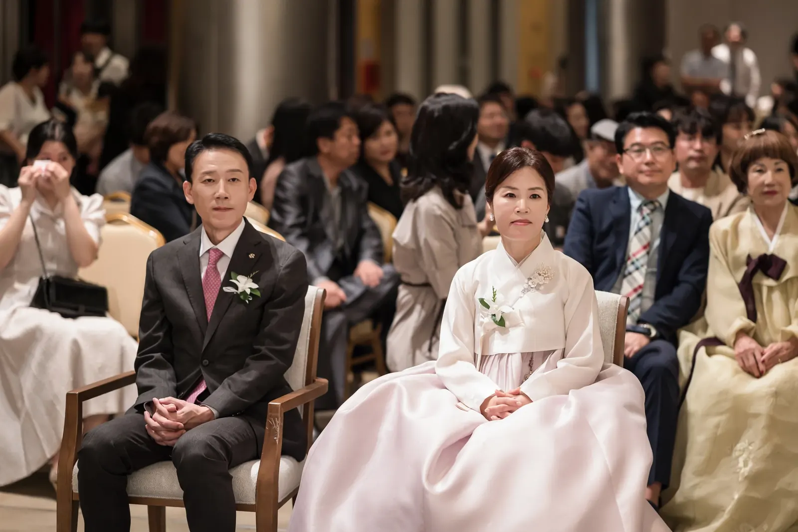 혼주석 가족사진 — 서울 포스코컨벤션 하객석 맨 앞줄에 앉아 예식을 지켜보는 정장 차림 아버지와 한복 입은 어머니