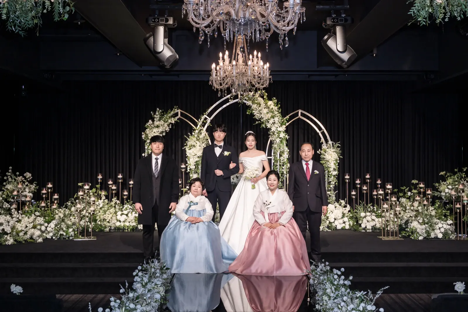 결혼식 가족사진 상담 — 서울 강남구 세인트메리엘 어두운 홀 샹들리에 아래 앉은 한복 혼주와 뒤에 선 가족, 신랑 신부