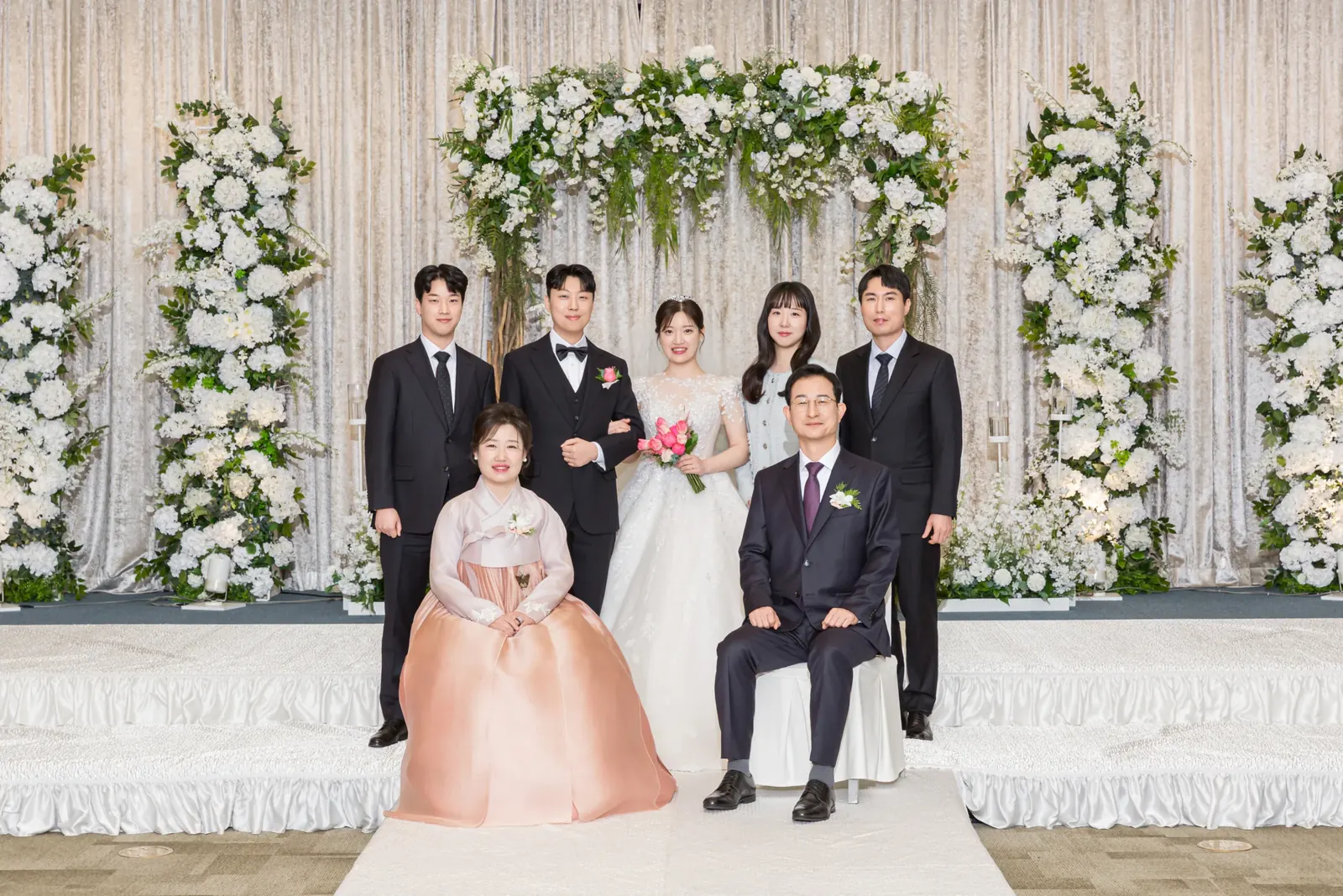 원판 가족사진 예시 — 서울 강동구 삼성E&A 꽃 아치 단상 앞에서 앉고 선 양가 가족과 신랑 신부