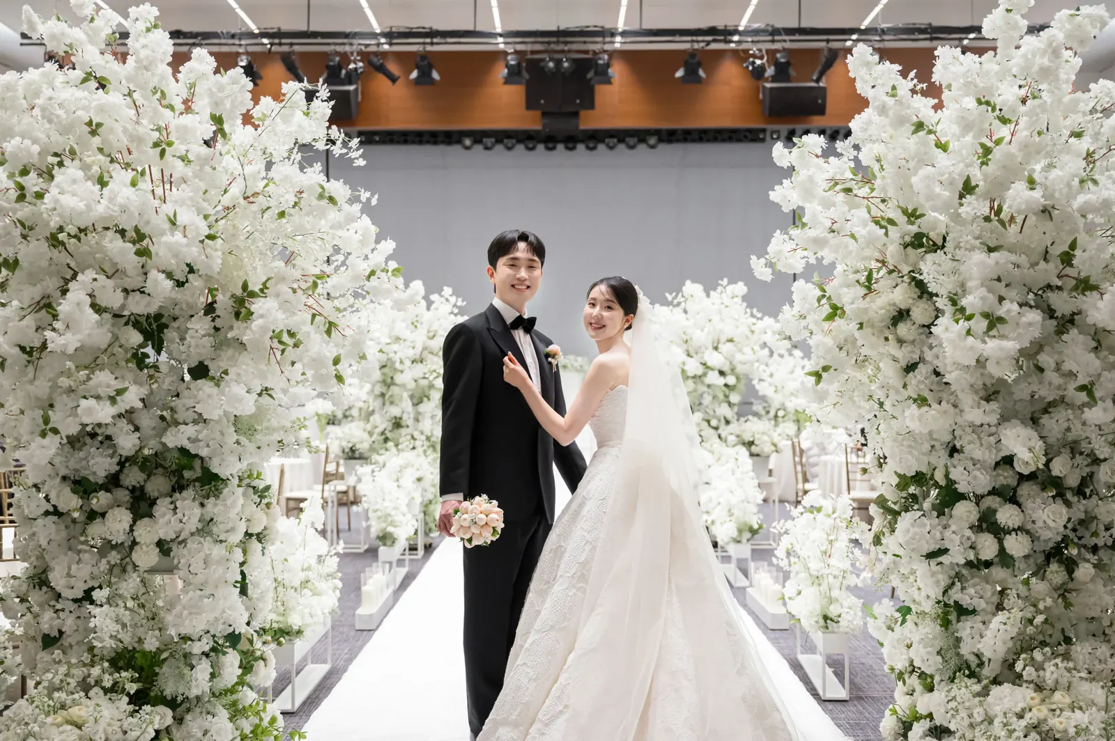 삼성서초사옥 결혼식사진 — 양옆 흰 꽃나무 장식 사이 버진로드에서 신부 어깨를 감싼 신랑