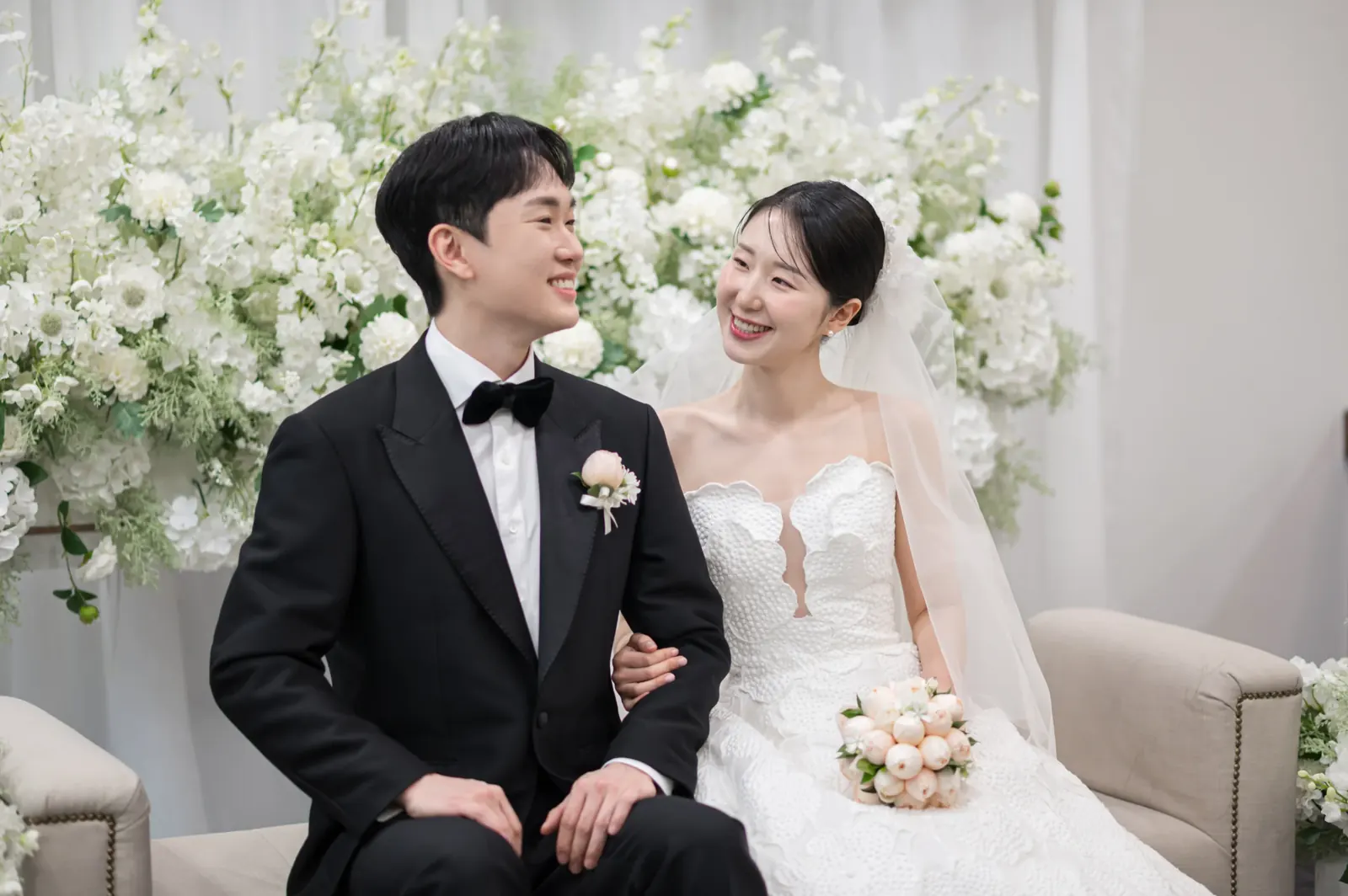 자연스러운 결혼식사진 예시 — 서울 서초구 삼성서초사옥 흰 꽃장식 앞 소파에 앉아 서로를 바라보며 웃는 신랑 신부