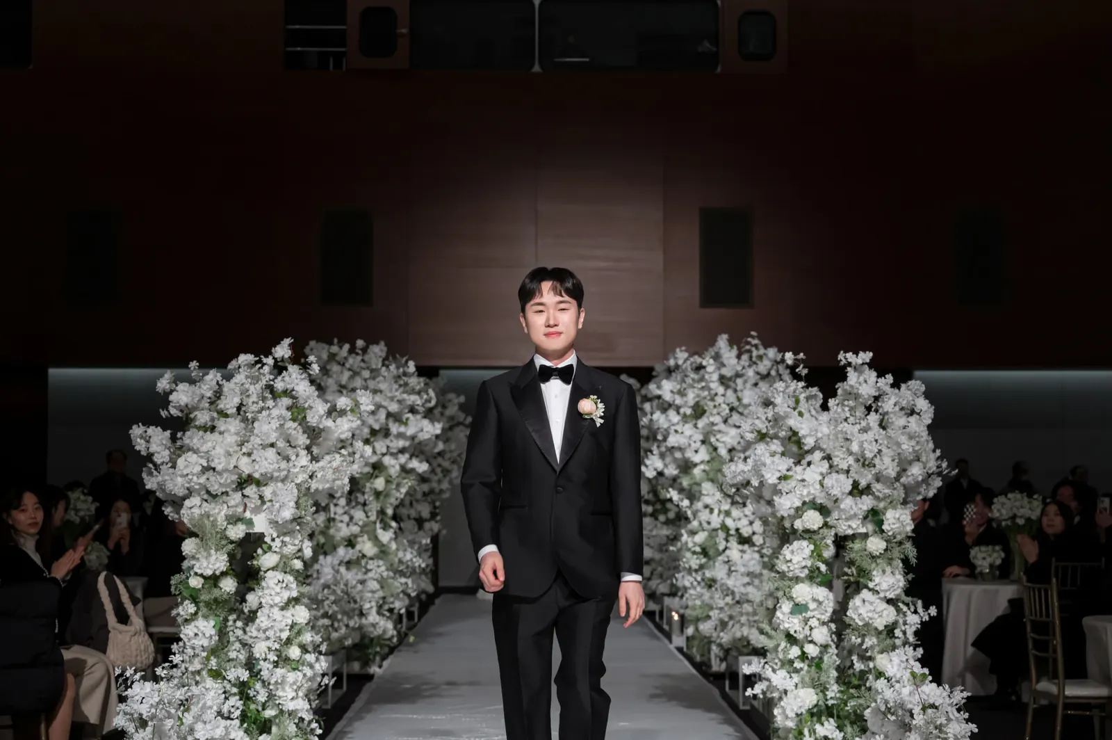 삼성서초사옥 결혼식사진 — 어두운 홀 흰 꽃나무 사이 버진로드로 입장하는 신랑