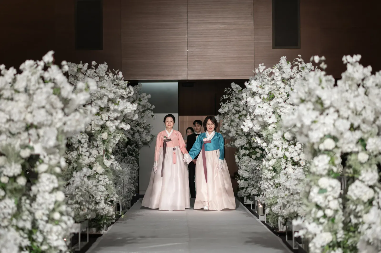 삼성서초사옥 결혼식사진 — 흰 꽃나무 버진로드로 함께 입장하는 분홍·청록 한복의 양가 어머니