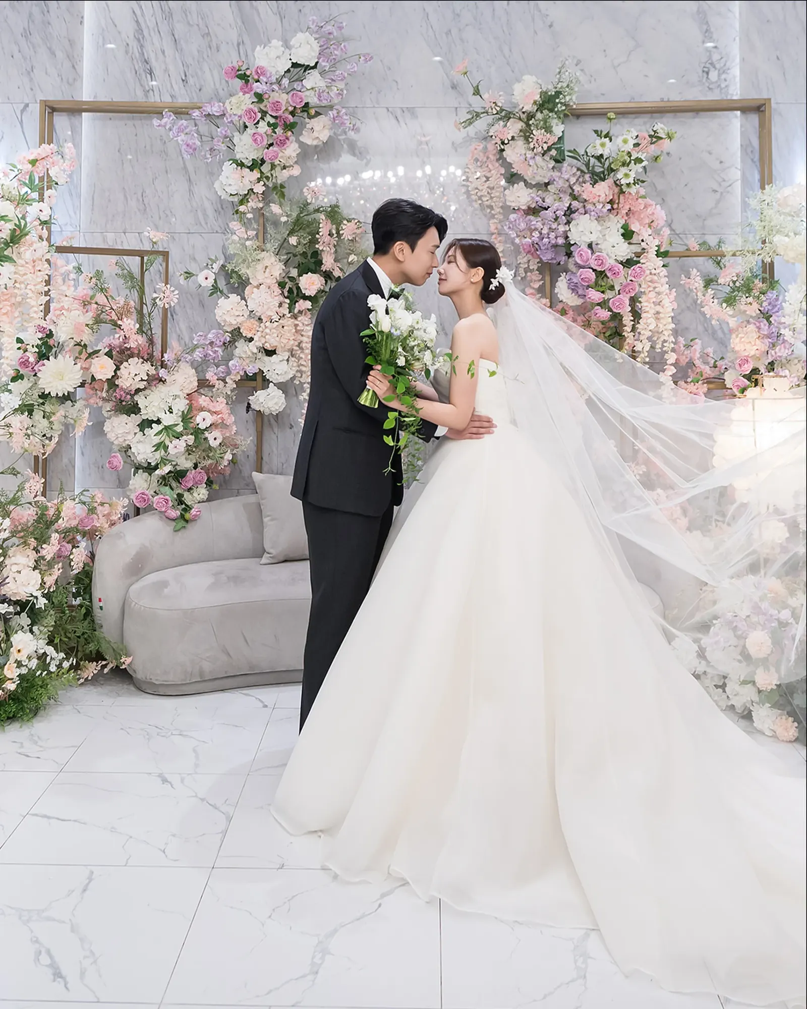 예식 촬영 문의 — 서울 신도림 웨딩시티 파스텔 꽃장식과 대리석 벽 앞에서 베일을 날리며 입맞춤하는 신랑 신부